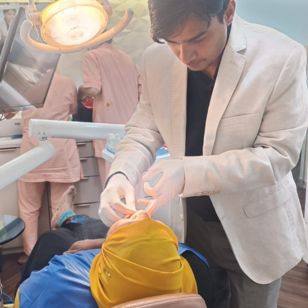 Male dentist in a beige blazer performing a dental procedure on a patient wearing a bright yellow headscarf in a clinic.