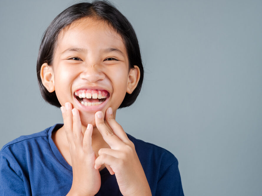 Cheerful young girl with short black hair in a blue shirt, smiling broadly with hands near her mouth against a gray-blue background.