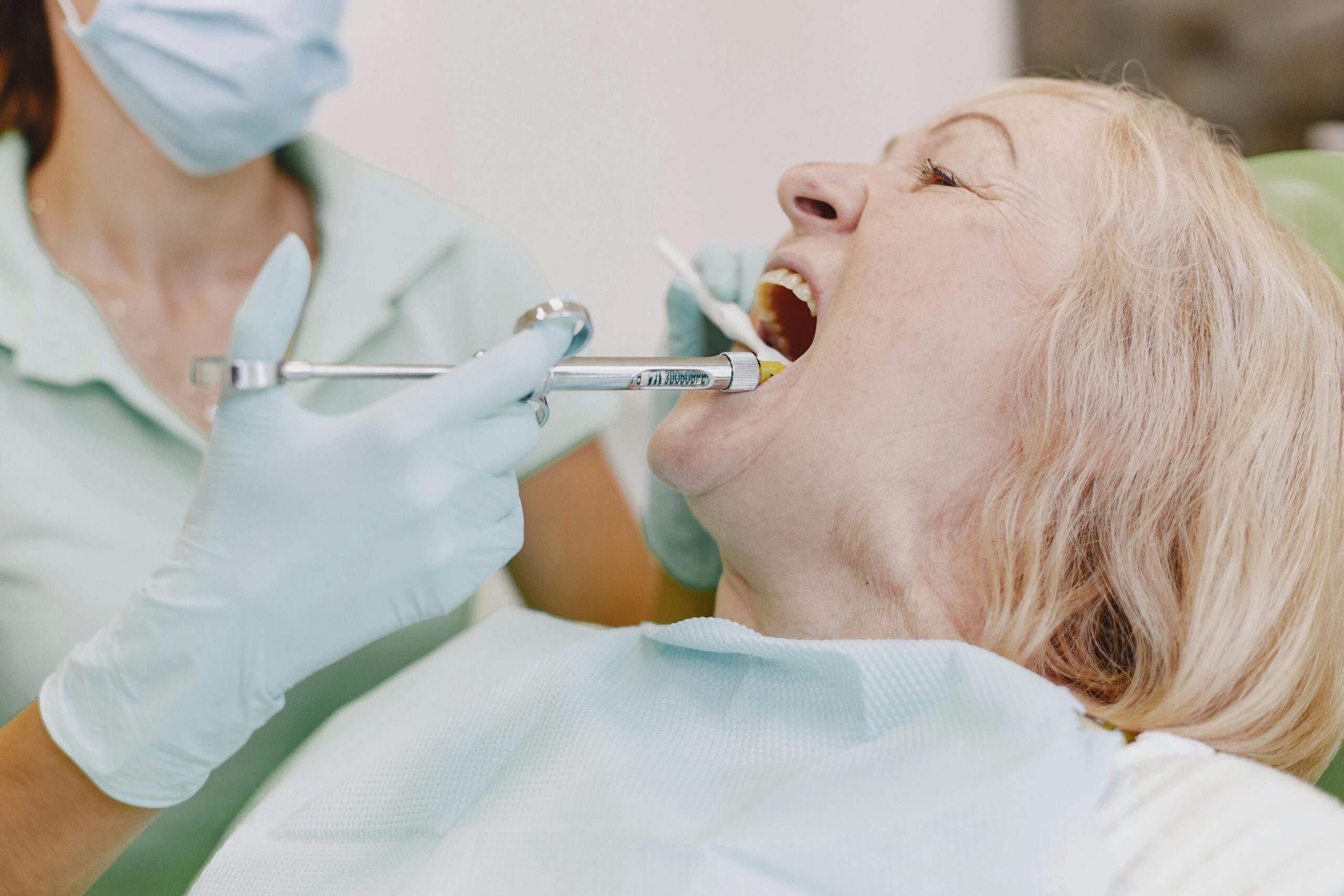 Dental professional wearing a mask and gloves performs a dental exam on a patient who is lying back in the chair, with a dental tool in the mouth.