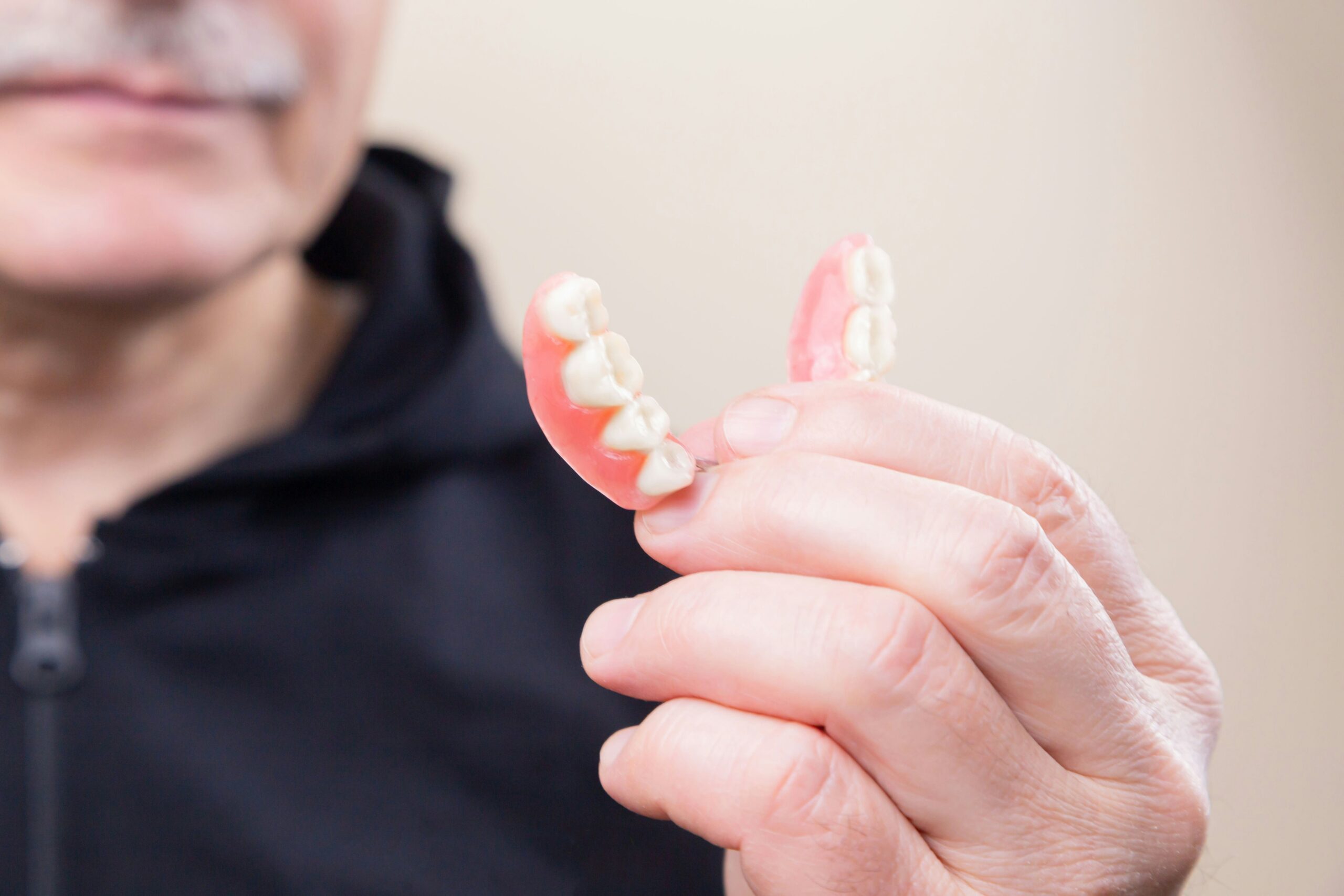Close-up of a pink partial denture with white teeth held between fingers, against a blurred background.