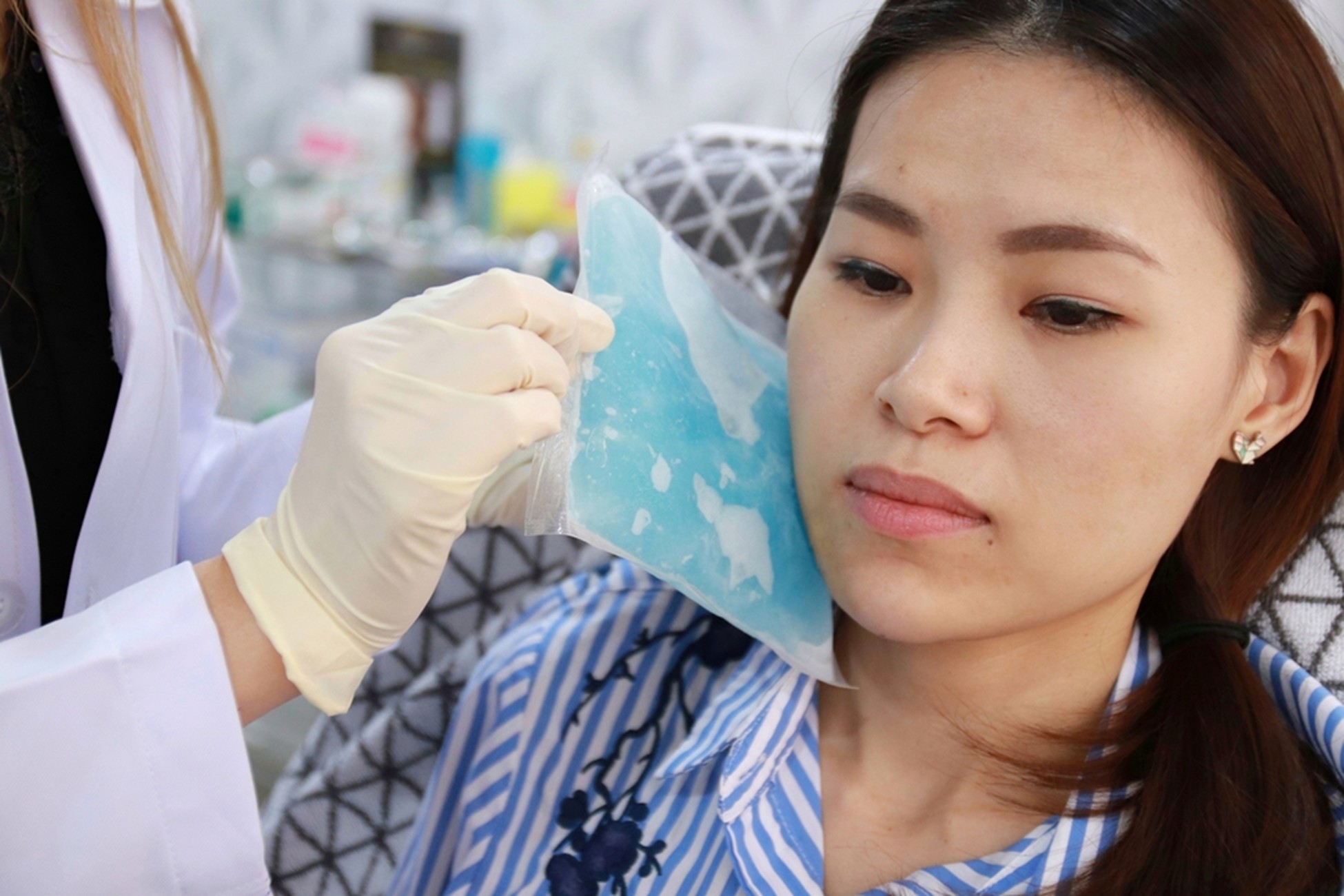 Healthcare worker wearing gloves applies a blue surgical mask to a woman's face, in a clinical setting.