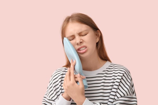 Picture2 Woman in a striped shirt presses a blue ice pack to her cheek, eyes closed in discomfort.