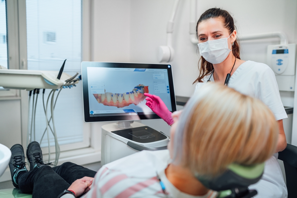 Dentist,Female,Doctor,In,Uniform,Showing,Scan,Of,Intraoral,3d Dentist in a mask points to a 3D dental model on a monitor while a patient sits in a dental chair.